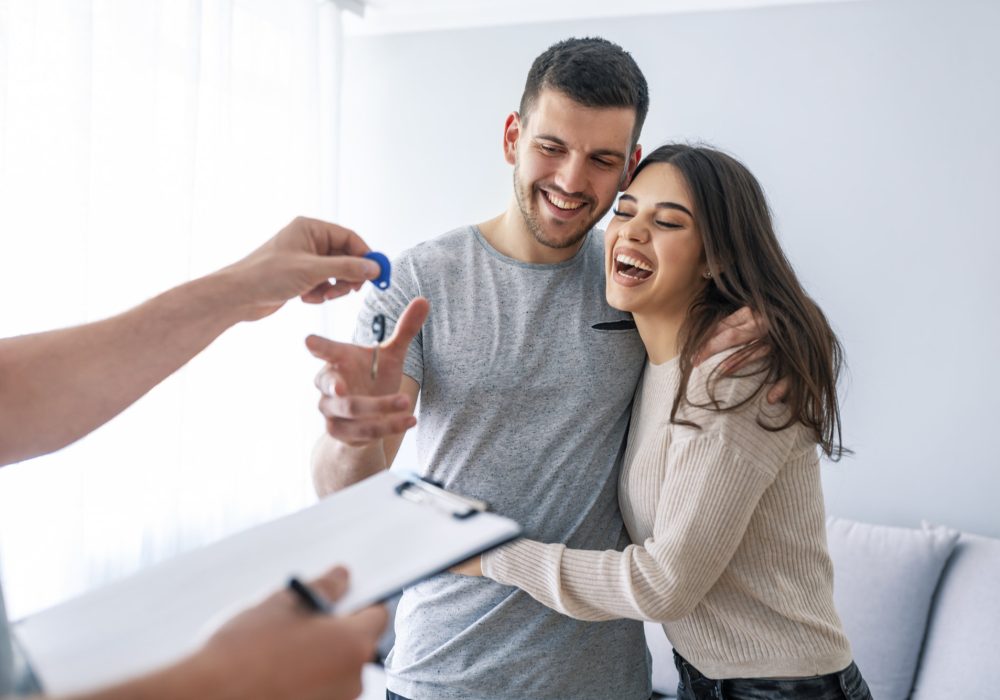 Happy couple is taking keys from their new house from broker and smiling. Hands of estate agent giving keys to the couple. The agent handed the keys a young couple